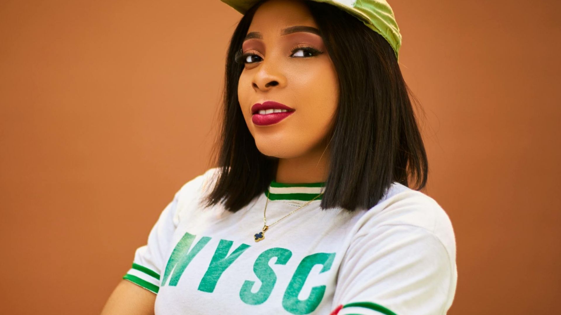 Woman wearing an NYSC white t-shirt with green lettering and a green cap, posing against an orange background.