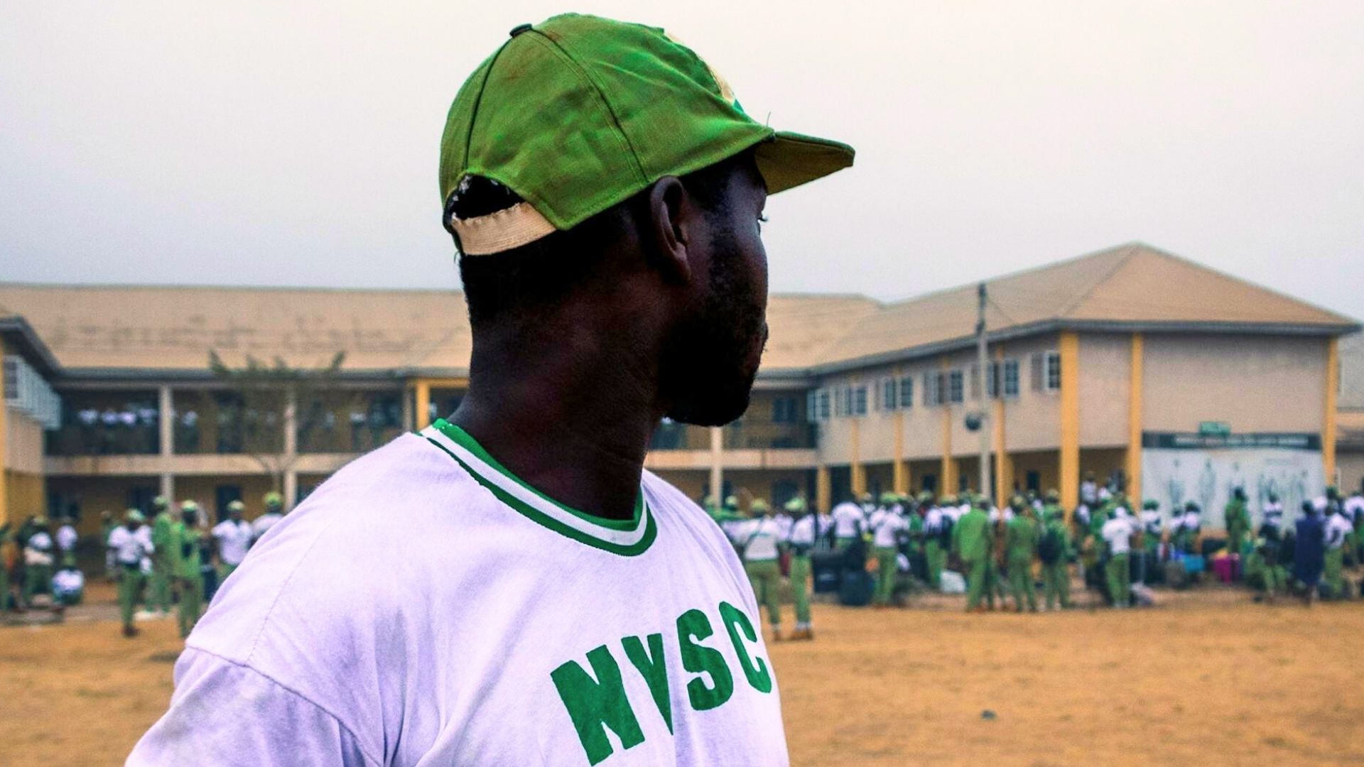 Person wearing a NYSC uniform, standing in front of a school building, with other people in similar attire in the background.
