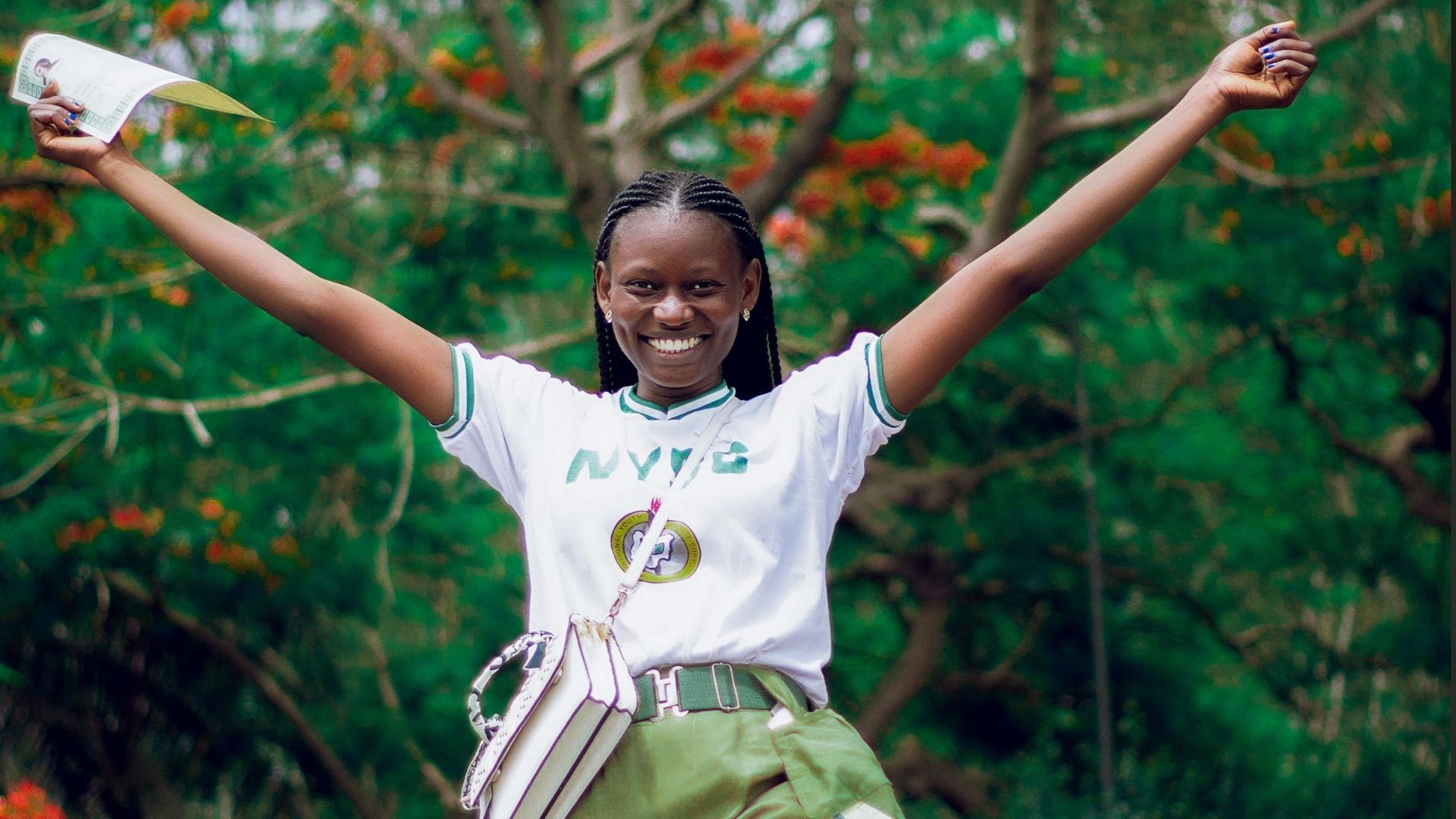 Smiling young woman with arms raised, holding a document, wearing a white shirt with green accents and olive green pants, outdoors.