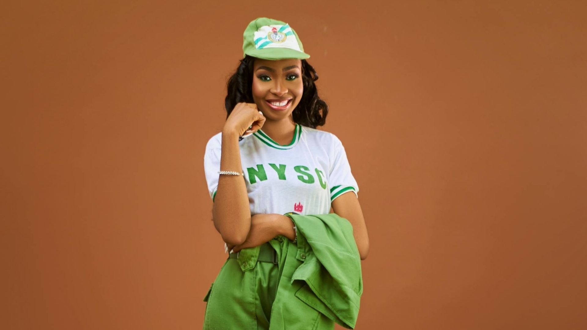 Woman wearing NYSC uniform, white t-shirt with green accents, and a green jacket, against a terracotta background.